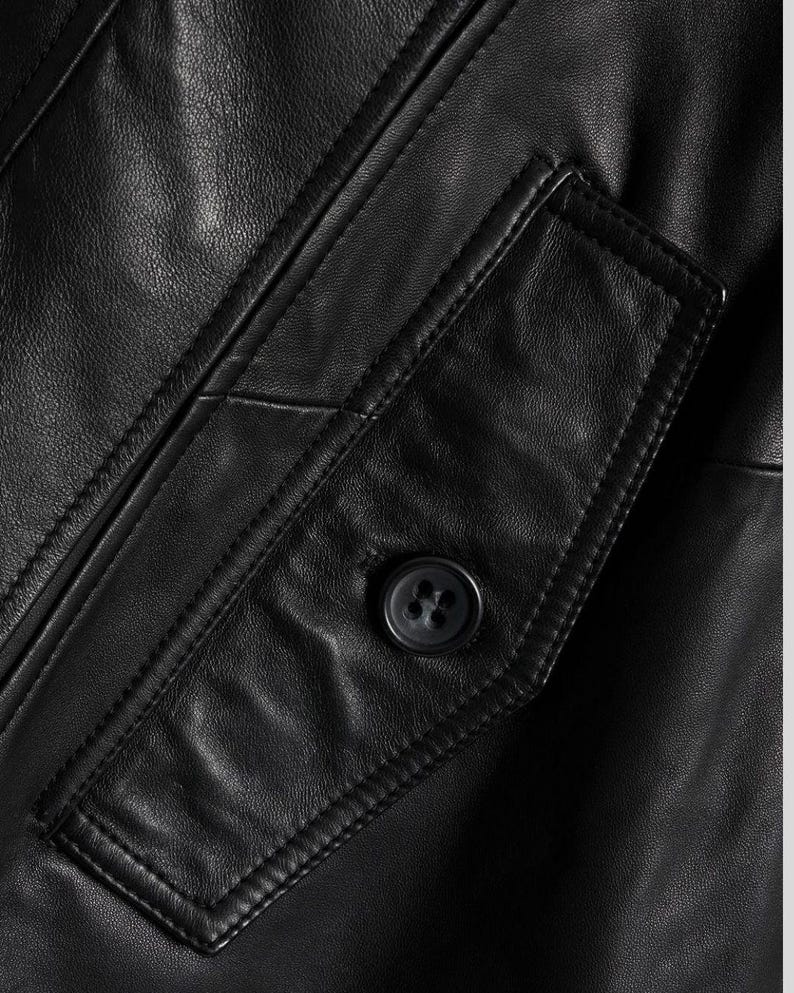 May include: Close-up of a black leather jacket featuring a buttoned pocket. The jacket has visible stitching and a textured surface. The button is dark and round, contrasting with the smooth leather.