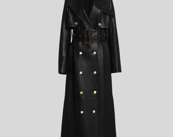May include: A long, black leather trench coat with a double-breasted design. The coat features a belt with a rectangular buckle and gold-colored buttons. The coat has a classic trench coat collar and shoulder epaulettes.