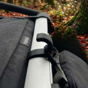 Puede incluir: Primer plano de un cochecito negro y gris con un marco plateado. El cochecito tiene un gancho negro y una correa gris. La marca "THULE" es visible. El fondo está borroso con follaje otoñal.