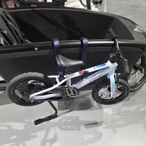Peut inclure: Un vélo enfant bleu clair attaché à un vélo cargo noir. Le petit vélo a des pneus noirs et une selle noire. Le vélo cargo a un compartiment de rangement noir et un siège noir pour enfant. Les vélos sont sur une surface grise.