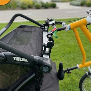 May include: A black Thule bike trailer with a yellow and white bicycle attached. The trailer has a mesh cover and a black handle. The bicycle has a yellow frame and white handlebars. The scene is set on green grass.