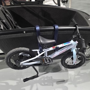 Puede incluir: Una bicicleta de carga negra con un contenedor de almacenamiento grande y profundo con correas de seguridad internas, junto con una bicicleta pequeña para niños de color azul claro sujeta al lateral del marco de la caja de carga.