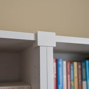 以下が含まれることがあります： 白の本棚。棚とカラフルな本の一部が見えます。本棚は、シンプルで幾何学的な構造を持つ、クリーンでモダンなデザインです。棚には、さまざまなサイズと色の本が並んでいます。