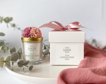 May include: A clear glass candle jar with a gold rim and a small white box, both with the text "YOUR LOGO HERE" and "YOUR CUSTOM TEXT HERE". The candle jar is topped with a pink and peach flower arrangement. A pink ribbon is tied on top of the box.