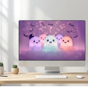 May include: A desktop computer shows a Halloween image of five smiling, glowing ghosts in purple, white, and orange. The ghosts are set against a bokeh background with bat silhouettes. The computer sits on a wooden desk with a keyboard, mouse, and small potted plants.