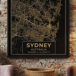 May include: A framed map of Sydney, Australia, with a gold-colored city layout on a black background. The city name and coordinates are displayed in a rectangular box at the bottom. The map is hanging on a brick wall.