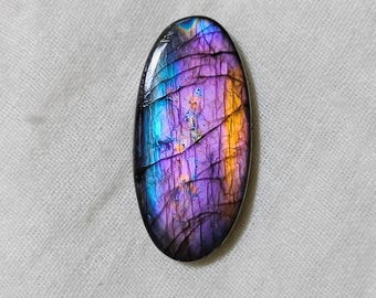 May include: Oval-shaped labradorite gemstone with a vibrant display of colors. The stone features a mix of blues, purples, and oranges, with a crackled pattern across its surface. The gemstone is set against a white fabric background.