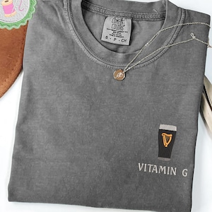 May include: A gray Comfort Colors t-shirt with a small embroidered design of a pint of stout with a harp and the text "VITAMIN G". A silver necklace with a pendant is draped on the shirt. The shirt is folded.