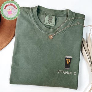 May include: Green Comfort Colors t-shirt with an embroidered pint of beer and the text "Vitamin G." A gold necklace with a pendant rests on the shirt. The shirt is folded on a white surface.