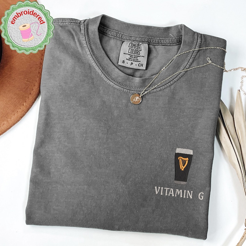 May include: A gray Comfort Colors t-shirt with a small embroidered design of a pint of stout with a harp and the text "VITAMIN G". A silver necklace with a pendant is draped on the shirt. The shirt is folded.