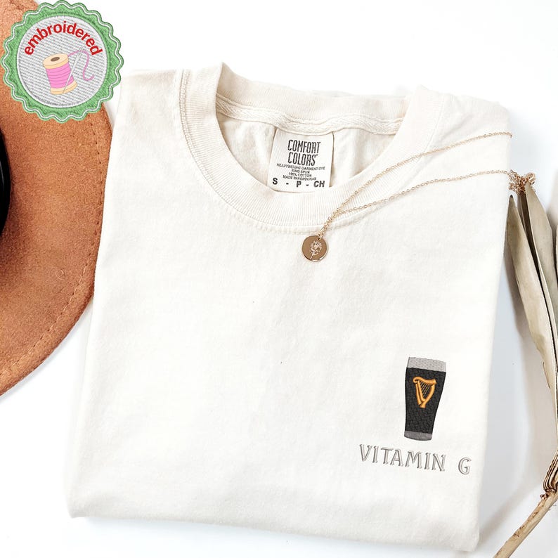 May include: Cream-colored t-shirt with a small embroidered design of a pint of stout with a gold harp and the text "Vitamin G". A gold necklace with a pendant rests on the shirt. The shirt has a Comfort Colors tag.