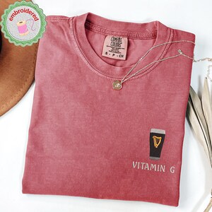 May include: A dusty rose Comfort Colors t-shirt with a small embroidered design of a pint of stout and the words "Vitamin G". A silver necklace with a small pendant is draped on the shirt. The shirt is folded.