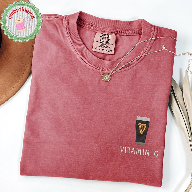 May include: A dusty rose Comfort Colors t-shirt with a small embroidered design of a pint of stout and the words "Vitamin G". A silver necklace with a small pendant is draped on the shirt. The shirt is folded.