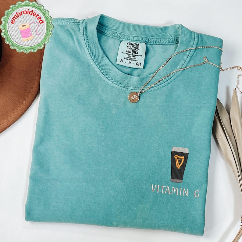 May include: A teal Comfort Colors t-shirt with an embroidered design of a pint of stout and the text "Vitamin G". A gold necklace with a pendant rests on the shirt. The shirt is folded and has a small tag.