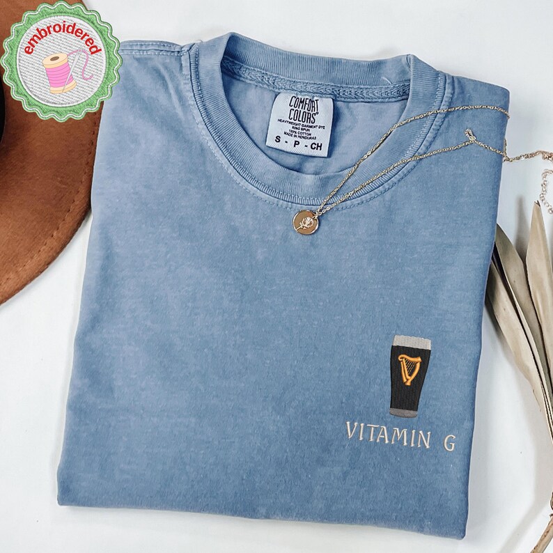 May include: A blue Comfort Colors t-shirt with an embroidered design of a pint of stout and the words "Vitamin G". A gold necklace with a pendant is draped across the shirt. The shirt has a small tag that says "S - P - CH".