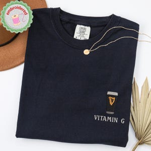 May include: Navy blue t-shirt with an embroidered design of a pint of stout and the text "Vitamin G". A gold necklace with a small pendant rests on the shirt. The shirt has a Comfort Colors tag.