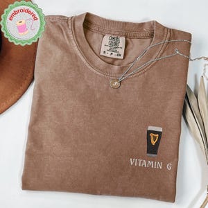 May include: Brown t-shirt with a crew neck and short sleeves. The shirt features an embroidered design of a pint of beer with the text "Vitamin G". A silver necklace with a pendant is draped across the shirt.