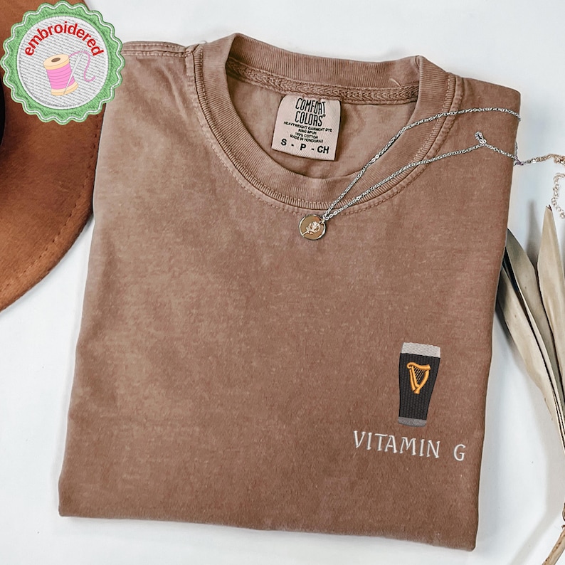 May include: Brown t-shirt with a crew neck and short sleeves. The shirt features an embroidered design of a pint of beer with the text "Vitamin G". A silver necklace with a pendant is draped across the shirt.