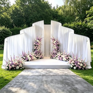 May include: A large, curved white fabric backdrop featuring a central opening, set upon a white platform. The structure is decorated with clusters of pink, purple, and white flowers, including roses and tall spikes, arranged along the base.