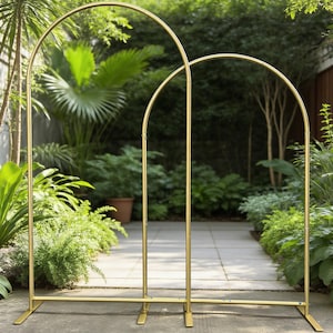 May include: Two gold metal arch structures, ideal for outdoor events, stand on a concrete surface. The arches have a rounded top and are supported by vertical poles and rectangular bases. Lush greenery forms the backdrop.