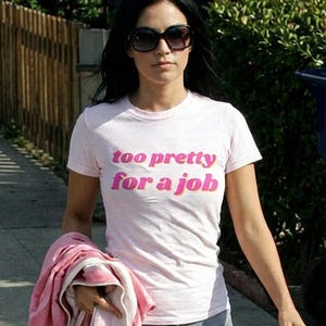 May include: A light pink t-shirt with the text "too pretty for a job" in pink lettering. The person is wearing grey shorts and sunglasses, holding a pink towel. The image is taken outdoors on a sunny day.