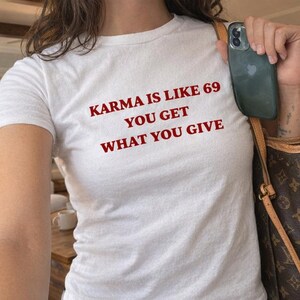 Puede incluir: Una camiseta blanca de manga corta con el texto rojo KARMA IS LIKE 69 YOU GET WHAT YOU GIVE impreso en el pecho. La prenda se combina con vaqueros azules y un bolso marrón con estampado, mientras se sostiene un smartphone oscuro en la mano.
