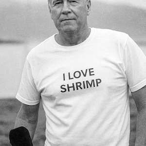 May include: A monochrome image of a person wearing a white t-shirt. The t-shirt has the words "I LOVE SHRIMP" printed in black. The shirt is a classic crew neck design.