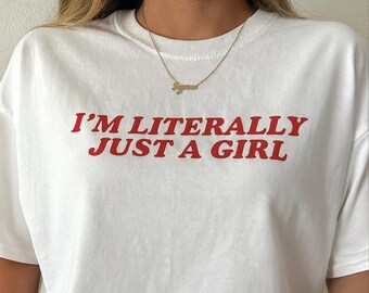Op de afbeelding: Wit T-shirt met de rode tekst "I'M LITERALLY JUST A GIRL". Een gouden ketting met een naamhanger wordt gedragen. Het shirt is een casual kledingstuk voor dagelijks gebruik.