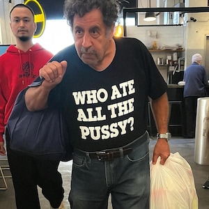 May include: A black t-shirt with the text "WHO ATE ALL THE PUSSY?" in white block letters. The person is wearing blue jeans and a watch. A person in a red hoodie is in the background.