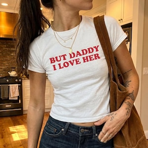 Puede incluir: Camiseta blanca con el texto rojo "BUT DADDY I LOVE HER". La camiseta se combina con vaqueros azul oscuro y un bolso tote de ante marrón. La persona lleva collares dorados.
