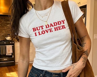Op de afbeelding: Wit t-shirt met de rode tekst "BUT DADDY I LOVE HER". Het shirt wordt gecombineerd met een donkerblauwe jeans en een bruine suède tote bag. De persoon draagt gouden kettingen.