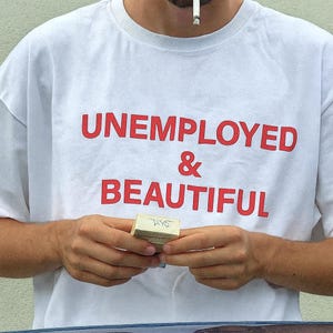 May include: White t-shirt with the red text "UNEMPLOYED & BEAUTIFUL". The person is holding a small, rectangular object. A lit cigarette is in the person's mouth.