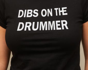 Könnte beinhalten: Schwarzes Crop-Top mit dem weißen Text "DIBS ON THE DRUMMER". Das Shirt hat einen Rundhalsausschnitt und kurze Ärmel. Der Text ist zentriert und in einer fetten, serifenlosen Schrift.