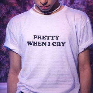 May include: A white short-sleeved t-shirt featuring the black text PRETTY WHEN I CRY printed on the chest. The shirt is worn with a white pearl bead necklace that sits around the neck. The fabric is plain white with a standard crew neckline.