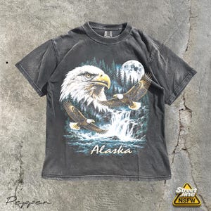Op de afbeelding: Een antracietgrijs t-shirt met korte mouwen en een grafische print van twee Amerikaanse zeearenden, een waterval, een bos en een volle maan. Het woord Alaska staat in een lichtgekleurd cursief lettertype onderaan het ontwerp.