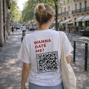 Op de afbeelding: Een wit T-shirt met korte mouwen en de rode tekst WANNA DATE ME? gedrukt boven een grote zwart-witte QR-code afbeelding. Het QR-code ontwerp bevat drie kleine hartvormige iconen die in de hoeken van het vierkante kader zijn geplaatst.