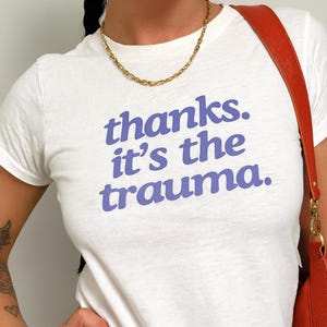 Puede incluir: Camiseta blanca con el texto "thanks. it's the trauma." en letras azules. También se ven un collar de cadena dorada y un bolso de hombro rojo.