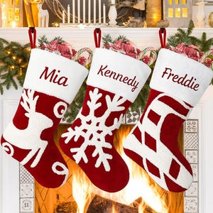May include: Three festive Christmas stockings in red and white hang above a fireplace. Each stocking has a white cuff with a name embroidered on it: Mia, Kennedy, and Freddie. The stockings feature different white patterns: a reindeer, a snowflake, and a diamond design.