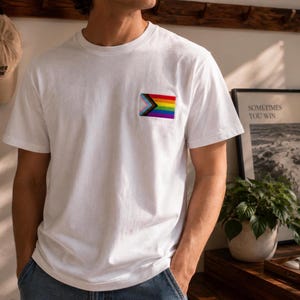 Puede incluir: Camiseta blanca con un pequeño parche de la bandera del Orgullo en el pecho. La bandera presenta un diseño de arcoíris con un galón rosa, celeste, blanco, marrón y negro. La camiseta es de manga corta y de material suave.