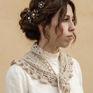 Puede incluir: Una mujer con cabello oscuro recogido, lleva un chal de ganchillo color crema y una blusa blanca. El chal tiene un borde festoneado y está atado en la parte delantera. Un clip floral adorna su cabello.