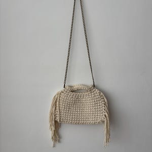 May include: A cream-coloured, crocheted handbag with a fringe detail. The bag is suspended by a dark metal chain. The bag has a rounded shape with a handle at the top.