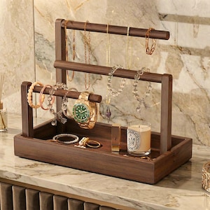 Puede incluir: Un organizador de joyas de madera oscura con múltiples niveles para collares, pulseras y anillos. Un reloj dorado, pulseras y una vela se exhiben en el organizador. El fondo presenta una pared similar al mármol.