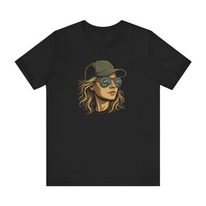 以下が含まれることがあります： 黒いTシャツ。緑の帽子とサングラスをかけた人物の漫画のグラフィック。サングラスは風景を映し出しています。その人は長く、波状のブロンドの髪をしています。