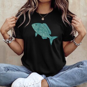 May include: Black t-shirt featuring a detailed teal fish design. The fish has intricate patterns and is positioned on the front of the shirt. The shirt is a classic crew neck style.