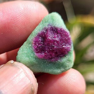 May include: A small, triangular gemstone with a deep magenta centre and a green matrix. The stone is held in a hand, with a blurred background, suggesting a natural setting.