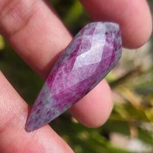 Könnte beinhalten: Ein facettierter, länglicher Edelstein in tiefem Magenta, der an den Rändern in Grau-Grün übergeht. Der Edelstein wird zwischen Fingern gehalten, wodurch seine polierten Facetten und natürlichen Farbvariationen hervorgehoben werden. Der Stein ist spitz zulaufend, mit einem spitzen Ende.