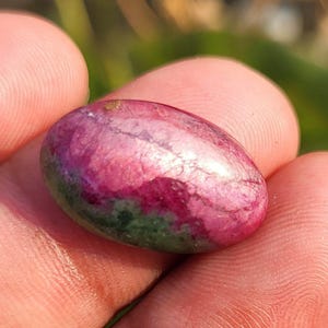 May include: An oval gemstone with a polished surface. The stone displays a mix of colours, including deep red, pink, and green hues. The gemstone is held in a person's hand, with a blurred background.