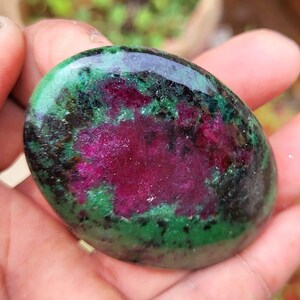 Puede incluir: Una gema pulida de forma ovalada con una vibrante mezcla de colores. La piedra muestra un centro magenta intenso, rodeado de áreas verdes y negras. La superficie es lisa y reflectante, resaltando los patrones naturales.