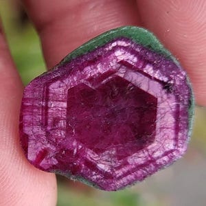 May include: A magenta-coloured gemstone with a green border. The stone is hexagonal, with visible internal structures and a polished surface. The gem's natural beauty is highlighted by its facets.