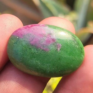 Puede incluir: Una gema pulida de forma ovalada, con una base verde vibrante y una prominente mancha de color rosa rojizo intenso. La piedra se sostiene en una mano, con un fondo natural borroso.